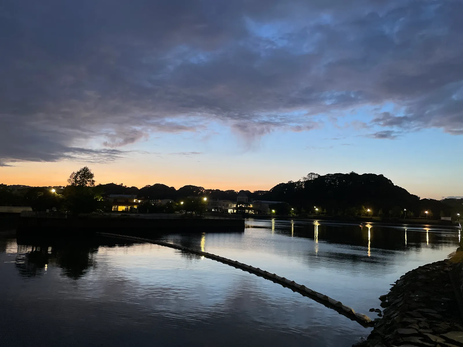 薄暮の水辺(入江または船溜まりのような静水面)を写した写真。対岸には低い建物群と街灯の明かりが並び、背後に木々の茂った丘陵のシルエットが広がる。水面には街灯やビル灯りが縦に反射している。手前には水面を横切るオイルフェンス状の浮きロープが写り、右手には石積みの護岸が見える。空はオレンジから紺色へのグラデーションで、夕暮れ時(または明け方薄明)の時間帯と推測される。季節感を示す要素(紅葉・新緑など)は乏しい。