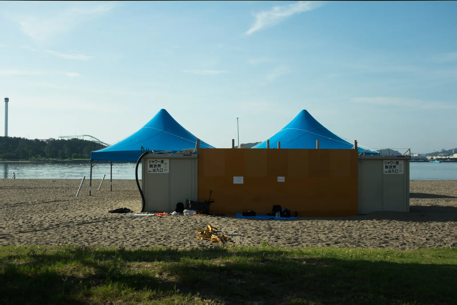 海水浴場に設置された仮設のシャワー室・脱衣所の青いテント2棟を正面から撮影。「シャワー室 脱衣所 出入口」の案内表示があり、男女別の入口が分かれている。背景には八景島シーパラダイスの展望タワーとジェットコースターの一部、対岸の山並みが見える。朝方の澄んだ青空で、海開き直後の設営状況がわかる資料的な写真。