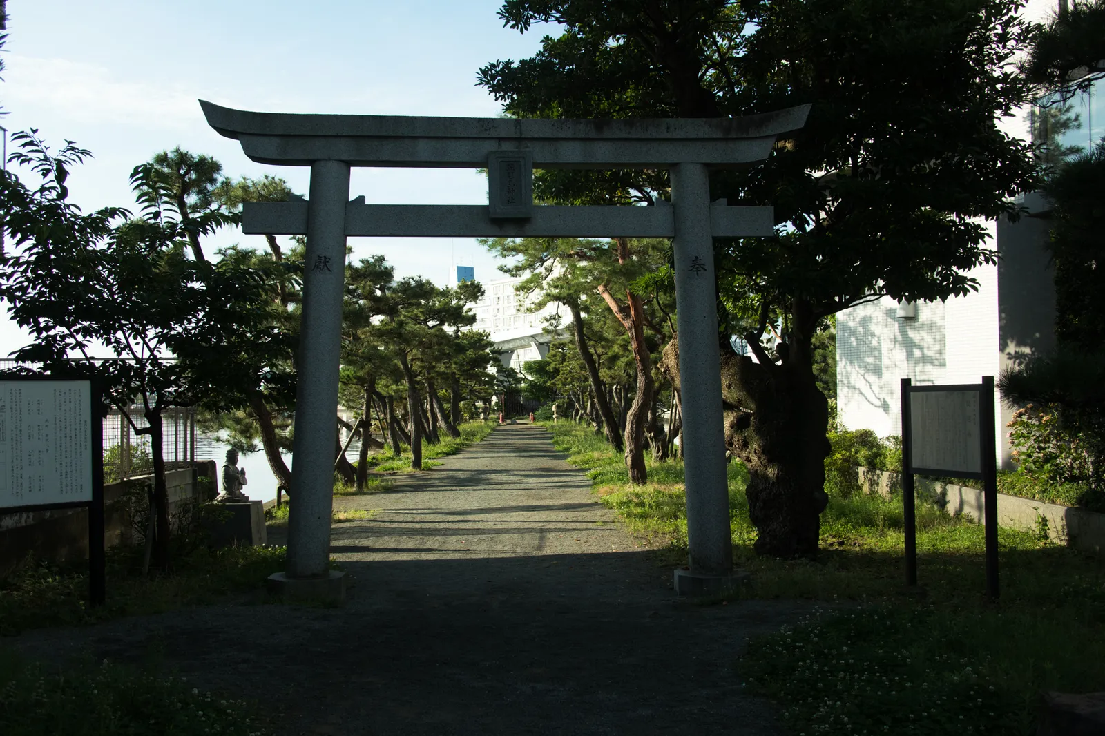 石造りの鳥居を正面から撮影。額束には「琵琶嶋神社」と読める文字が刻まれている。奥に松並木の参道が続き、左手に由緒書きの案内板と小さな石像、右手にも案内板が設置されている。