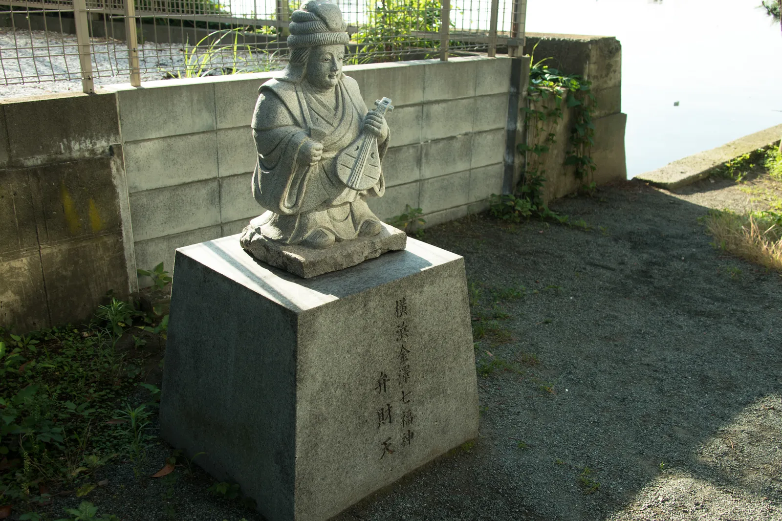 石造りの人物像(琵琶を抱えた弁財天像)。台座には「横濱金澤七福神 弁財天」の文字が刻まれている。ブロック塀と水辺を背景に、朝の斜光が当たっている。