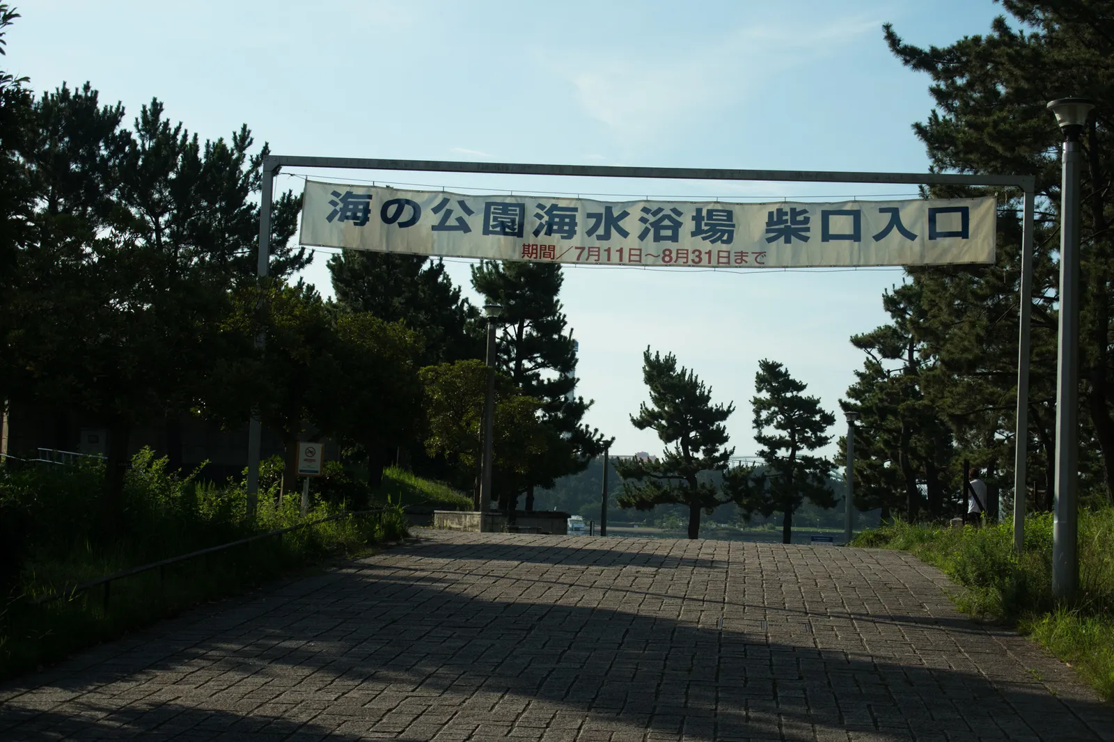 「海の公園海水浴場 柴口入口」と書かれた横断幕ゲート。期間は7月11日〜8月31日と記載されている。松林の中の石畳の坂道入口で、奥に人物が一人歩いている。海開き期間の案内を示す資料的な写真。