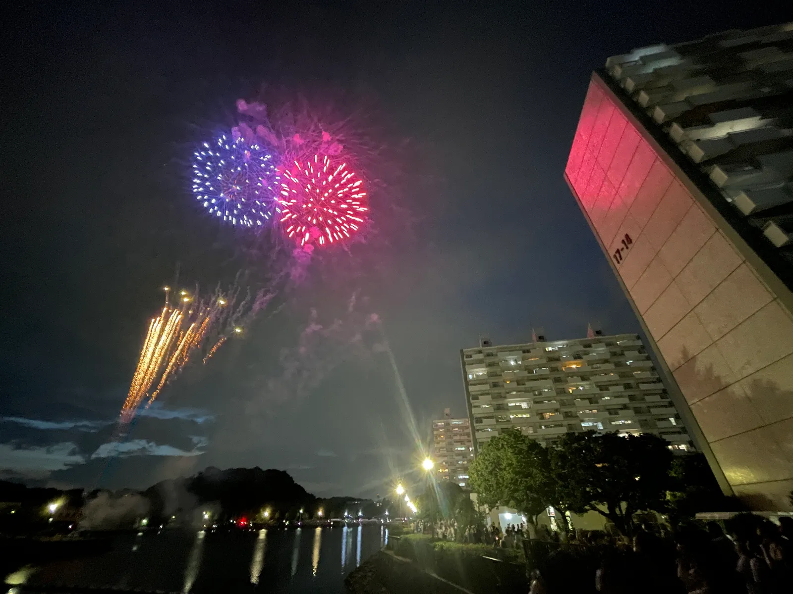 夜、船溜り付近から撮影した花火大会の写真。青色と赤紫(マゼンタ)の丸い花火が同時に開き、下方には金色の柳咲きの花火も重なっている。右側には花火の光でピンクに照らされた団地の壁面(「17-19」の表示)が写り、手前の水面や観覧する人々のシルエットも確認できる。